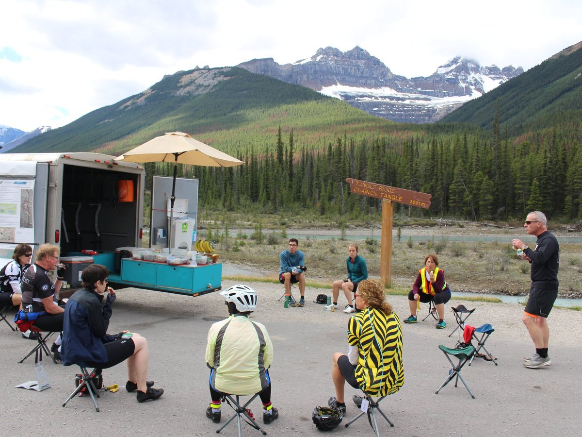 Riders relaxing at a rest stop beside the Mountain Madness van and trailer with mountains behind