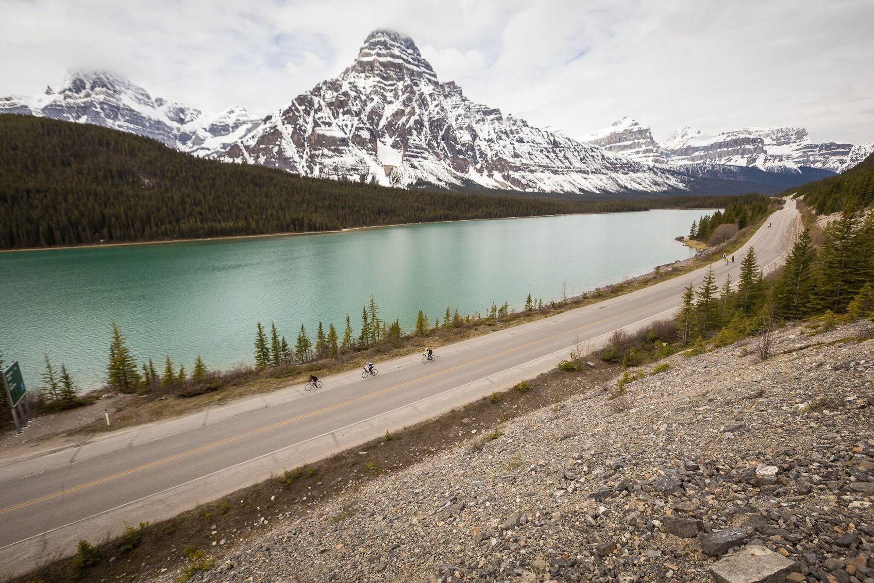 Riders on a lakeside road below snow-capped peaks