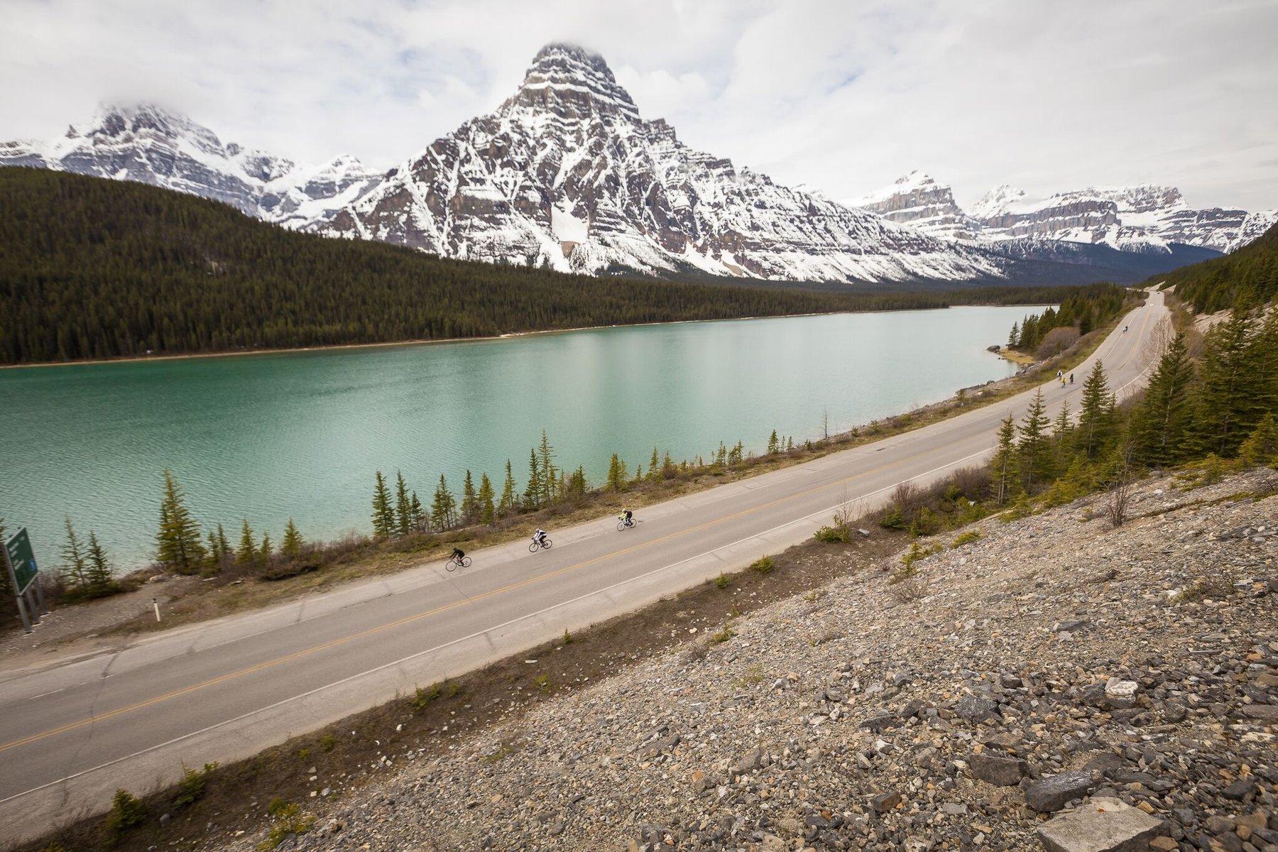 Riders on a lakeside road below snow-capped peaks